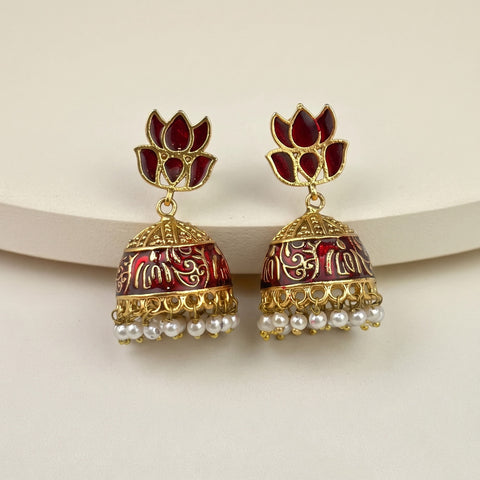 A pair of gold-plated Indian jhumka earrings with deep red floral studs and red-and-gold patterned bell-shaped drops, finished with small white pearl tassels, displayed on a curved cream-colored surface.