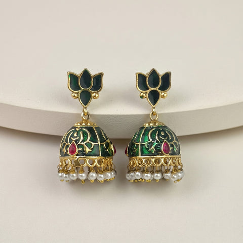 A pair of gold-plated Indian jhumka earrings with dark green enamel, floral engravings, small red teardrop accents, and dangling white pearl chains, displayed on a light beige surface.