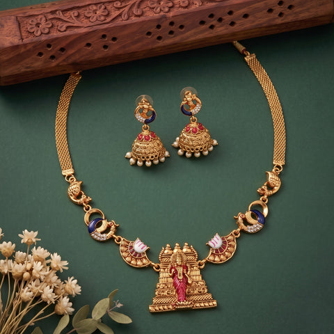 Gold-toned traditional necklace and matching earrings with red and blue stone accents, displayed on a dark green background with decorative dried flowers and a carved wooden prop.