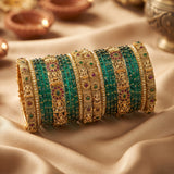 A stack of ornate Indian bangles (kara) featuring green beaded stripes, gold-plated metal bands with intricate filigree, and small red and green stone accents, displayed on draped beige fabric.
