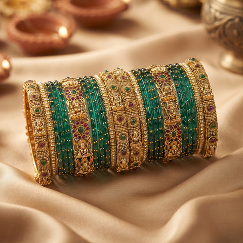 A stack of ornate Indian bangles (kara) featuring green beaded stripes, gold-plated metal bands with intricate filigree, and small red and green stone accents, displayed on draped beige fabric.