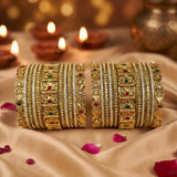 A set of ornate gold-toned Indian bangles (kara) decorated with intricate filigree, red and green stone accents, and silver-colored inlay, arranged on draped beige fabric with lit diya candles in the background.