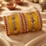 A stack of traditional Indian bangles, including yellow glass bangles, gold-plated bangles with red and green stones, and gold-plated bangles with silver stones, arranged on a beige satin fabric background with blurred diya lamps in the background.