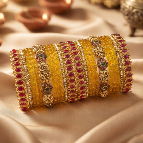 A stack of traditional Indian bangles, including yellow glass bangles, gold-plated bangles with red and green stones, and gold-plated bangles with silver stones, arranged on a beige satin fabric background with blurred diya lamps in the background.