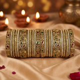 A stack of ornate gold-toned bangles with intricate filigree, beadwork, and small red and green stone accents, displayed on draped beige fabric with blurred lit diya candles in the background.