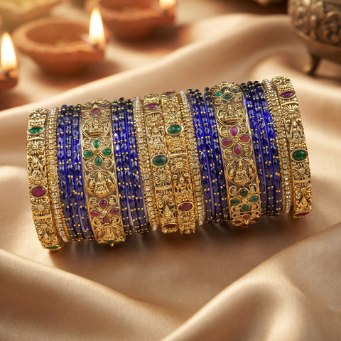 A set of ornate Indian bangles (kara) displayed on draped beige satin fabric with blurred diya candles in the background. The bangles feature gold-plated metal with intricate floral engravings, set with gold and silver crystals, and decorated with blue, green, and pink gemstones.