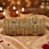 A stack of ornate gold bangles with intricate carvings, red and green stone accents, and silver-toned detailing, displayed on draped beige fabric with blurred lit diya candles in the background.