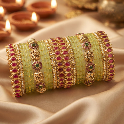 A stack of traditional Indian bangles, featuring light green beaded bands with intricate gold-plated borders, red stone accents, and small green and gold floral details, displayed on draped beige fabric with blurred diya candles in the background.