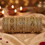 A stack of ornate Indian bangles (bangles) made of gold-plated metal, decorated with intricate motifs including Lord Ganesha, floral patterns, and small red and green stone accents. They are displayed on draped beige satin fabric with blurred diya candles in the background.