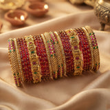 A stack of traditional Indian bangles (kara bracelets) made of gold-plated metal with red beaded stripes, pink stone accents, and green and red enamel details, displayed on a draped beige satin fabric.
