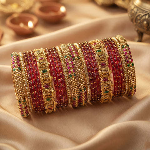 A stack of traditional Indian bangles (kara bracelets) made of gold-plated metal with red beaded stripes, pink stone accents, and green and red enamel details, displayed on a draped beige satin fabric.