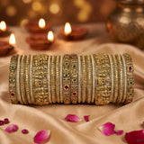 A stack of ornate gold-toned bangles with intricate floral and deity motifs, accented with small red and green stones, displayed on draped beige fabric with lit diya candles in the background.
