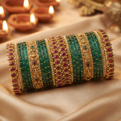 A set of traditional Indian bangles, including green stone bangles, gold-plated bangles with red stones, and gold-plated bangles with small clear stones, arranged on a beige satin fabric with blurred diya candles in the background.