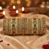 A set of ornate gold-toned Indian bangles (kara) decorated with intricate filigree, white stone pavés, and colorful green and red gemstone accents, displayed on draped beige fabric with blurred diya candles in the background.