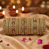 A stack of ornate gold bangles with intricate filigree, rhinestone embellishments, and small red and green stone accents, displayed on draped beige fabric with lit diya candles in the background.