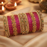 A set of traditional Indian bangles, including bright pink striped bangles and ornate gold-plated bangles with green and red stone accents, arranged on draped beige satin fabric with blurred diya lights in the background.