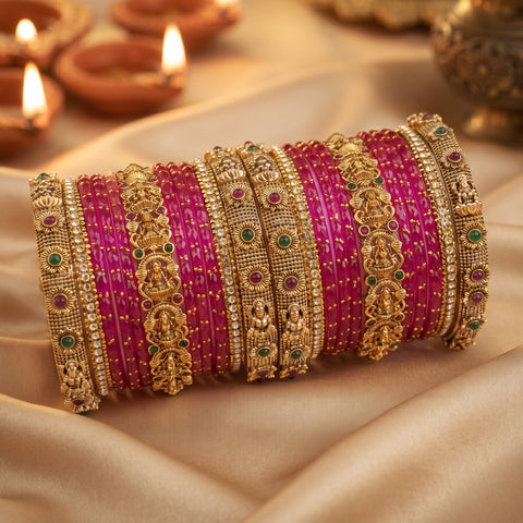 A set of traditional Indian bangles, including bright pink striped bangles and ornate gold-plated bangles with green and red stone accents, arranged on draped beige satin fabric with blurred diya lights in the background.