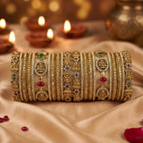 A stack of ornate gold bangles with intricate designs, set against a warm background with lit diya lamps and draped beige fabric.