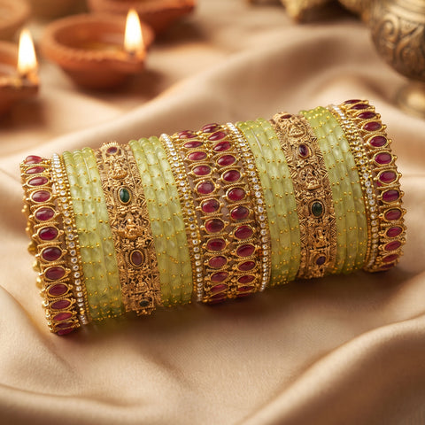 A stack of traditional Indian bangles, including light green stone-wrapped bangles, gold-plated bangles with red stones, and gold-plated bangles with white stones, arranged on draped beige fabric with blurred diya candles in the background.