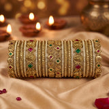 A stack of ornate gold-toned bangles with intricate silver-toned detailing, red and green stone accents, and engraved motifs, displayed on draped satin fabric with blurred lit diya candles in the background.