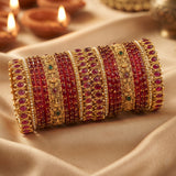 A stack of ornate Indian bangles (kara bracelets) featuring red stone inlays and gold-plated metal bands, displayed on draped beige fabric with blurred diya candles in the background.