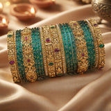 A stack of ornate Indian bangles, including turquoise green beaded bracelets and gold-plated filigree bangles with red and green stone accents, displayed on a beige satin fabric background.