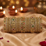 A stack of ornate gold bangles with intricate floral, geometric, and gemstone patterns, displayed on draped beige satin fabric with glowing diya candles in the background.