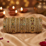 A stack of ornate gold-toned Indian bangles (bangles) decorated with intricate filigree, red and green stone accents, and traditional motifs, displayed on a draped beige satin fabric with blurred diya candles in the background.