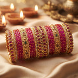 A stack of ornate Indian bangles (kara) made of gold-plated metal, decorated with rows of magenta-pink beads and set with red stones and small green accents, displayed on draped beige fabric with blurred diya candles in the background.