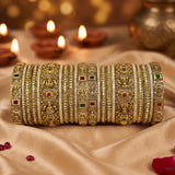A stack of ornate gold-toned bangles with intricate embossed motifs and small green and red stone accents, displayed on draped satin fabric with blurred lit candles in the background.