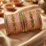 A stack of ornate Indian bangles (kara bracelets) featuring pink stone inlays, gold-plated borders, and red and green stone accents, displayed on draped satin fabric with blurred diya candles in the background.