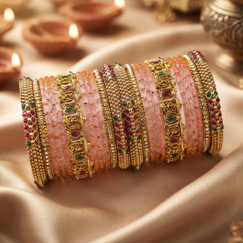 A stack of ornate Indian bangles (kara bracelets) featuring pink stone inlays, gold-plated borders, and red and green stone accents, displayed on draped satin fabric with blurred diya candles in the background.