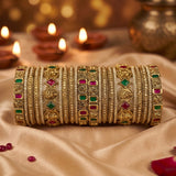 A stack of ornate gold bangles with red and green gemstone accents, displayed on draped satin fabric with blurred lit diya candles in the background.