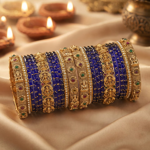 A stack of traditional Indian bangles, including royal blue beaded bangles and gold-plated bangles with deity motifs and small green and red stones, displayed on draped beige fabric with blurred diya candles in the background.