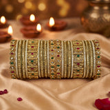 A stack of ornate gold bangles decorated with red, green, and white stones, displayed on draped beige satin fabric with lit diya candles in the background.