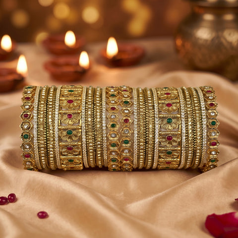 A stack of ornate gold bangles decorated with red, green, and white stones, displayed on draped beige satin fabric with lit diya candles in the background.