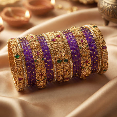 A stack of traditional Indian bangles, featuring purple beaded bands and ornate gold-plated filigree cuffs decorated with small red and green stones, displayed on draped beige satin fabric.