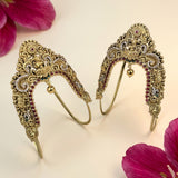 A pair of ornate gold-toned traditional Indian headwear pieces with intricate filigree work, red and white gemstone embellishments, and small hanging gold bells, displayed on a light cream background with pink flower petals in the corners.