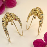 A pair of ornate gold-toned traditional Indian bridal headbands decorated with intricate filigree, small hanging bells, and colorful gemstones, displayed on a light cream background with pink flower petals.