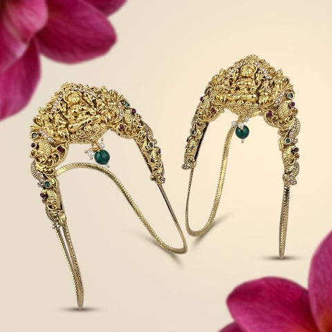 A pair of ornate gold filigree headbands decorated with small green and red gemstones, shown on a light beige background with pink flower petals in the corners.
