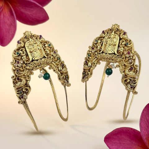 A pair of ornate gold-toned traditional Indian headpieces decorated with intricate filigree, small carved motifs, and colorful gemstones, displayed against a soft beige background with pink flower petals in the corners.