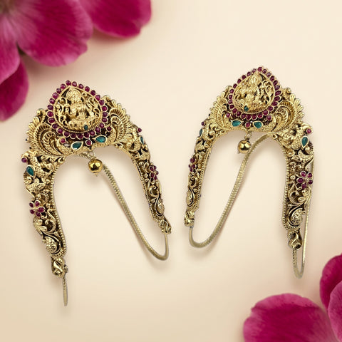 A pair of ornate gold-toned traditional Indian earrings with detailed filigree, red and green stone accents, and a central deity motif, displayed on a light cream background with pink flower petals.