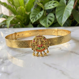 An ornate gold-toned traditional Indian bangle (kara) featuring a detailed central pendant with a deity motif and colorful enamel accents, displayed on a white marble surface with green plants in the background.