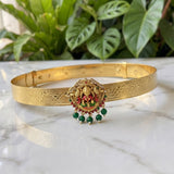 A gold-plated traditional Indian bangle (kara) featuring an ornate central Ganesha pendant decorated with red and green enamel and hanging green bead tassels, displayed on a white marble surface with green plants in the background.
