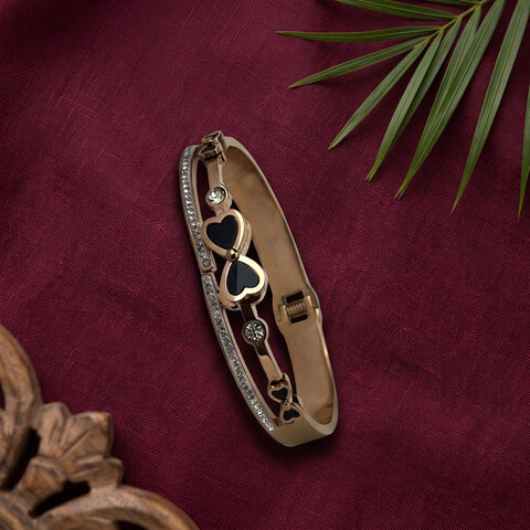 A gold-toned bangle bracelet with three interlocking heart charms, two of which are black, and a row of clear rhinestones along one side, displayed on a deep burgundy fabric background with decorative leaves and wood in the corners.