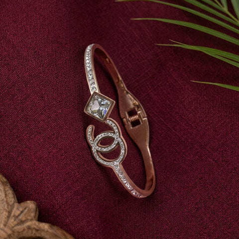 A rose gold bangle bracelet featuring a clear square crystal and a double-C logo design set with small crystals, displayed on a textured burgundy fabric background with decorative green leaves.
