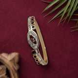 A gold-plated bangle bracelet with a central oval motif paved with clear crystals and black square accents, displayed on a textured dark red fabric background with decorative leaves and a wooden object.