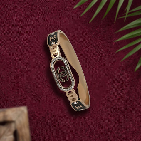 A gold-tone Chanel bangle bracelet with black enamel inserts, crystal-embellished oval links, and interlocking CC logos, displayed on a textured dark red fabric background with blurred green leaves.