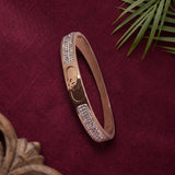A slim rose gold bangle bracelet with pavé crystal embellishments on the front, displayed on a textured dark red fabric background with decorative green leaves and a wooden frame.