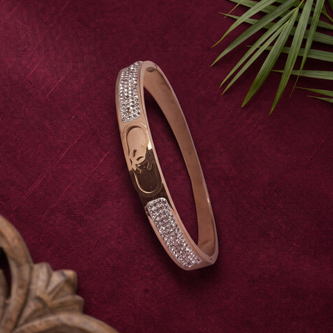 A slim rose gold bangle bracelet with pavé crystal embellishments on the front, displayed on a textured dark red fabric background with decorative green leaves and a wooden frame.